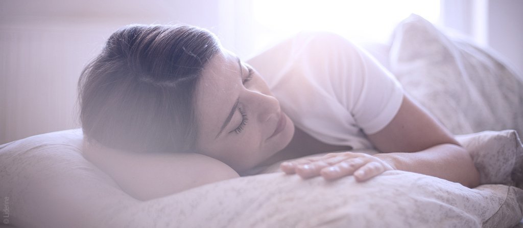 Foto: Eine Person liegt mit geschlossenen Augen im Bett. Durchs Fenster strahlt die Sonne herein.