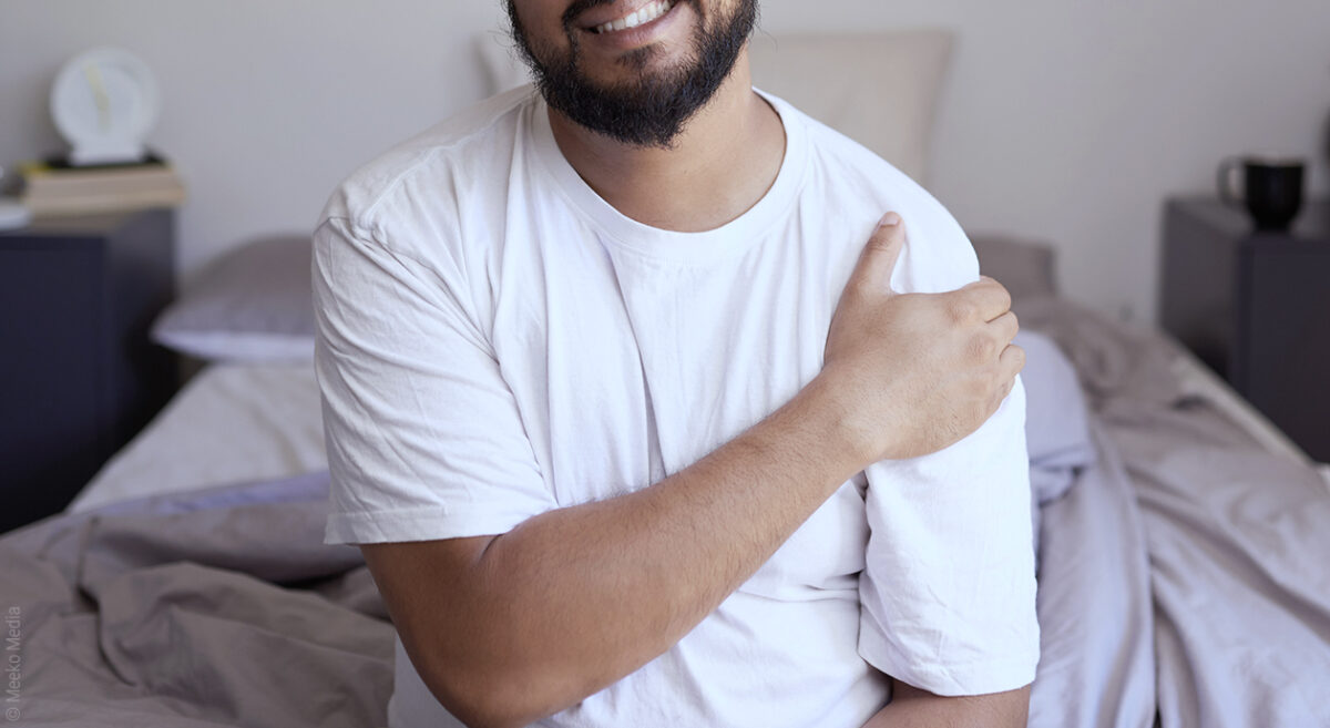 Foto: Eine Person sitzt auf der Bettkante und fasst sich mit schmerzverzerrtem Gesicht an die Schulter.