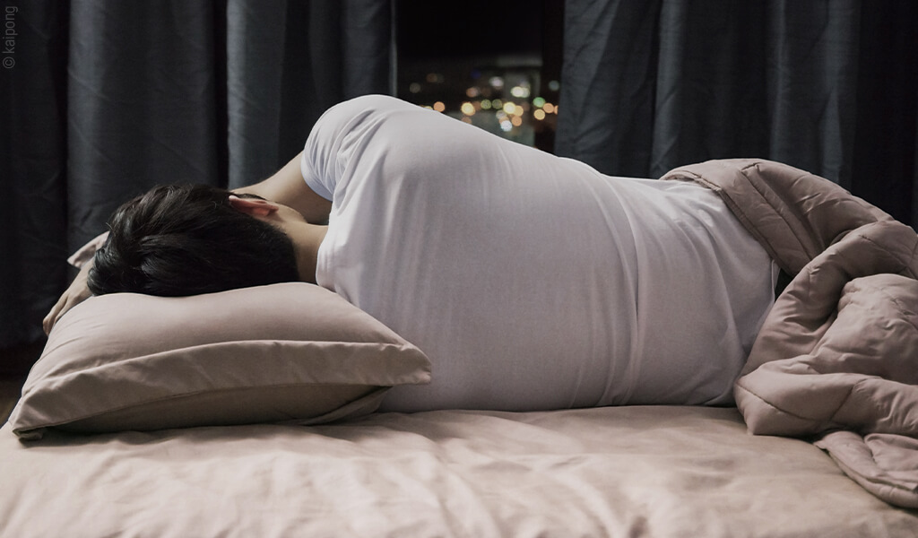 Person liegt als Seitenschläfer auf einem Kissen in einem Bett und schläft auf der Seite. Das Schlafzimmer ist abgedunkelt, im Hintergrund sind unscharfe Lichter zu sehen. Die Szene vermittelt eine ergonomische Schlafposition und ruhigen Schlafkomfort.