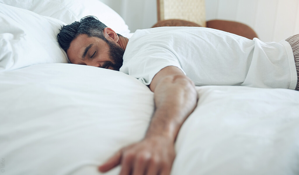 Person schläft entspannt in Bauchlage auf einem Bett mit weißer Bettwäsche. Ein Arm ist nach vorne ausgestreckt, der Kopf liegt seitlich auf einem Kissen. Die Aufnahme vermittelt Ruhe, Erholung und komfortablen Schlaf.